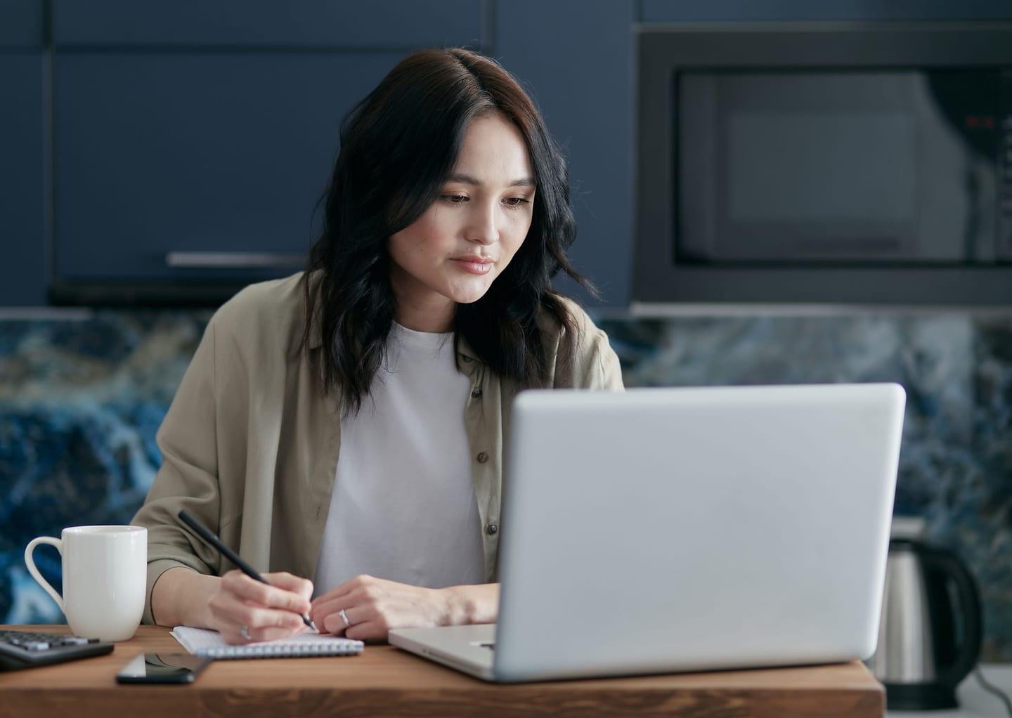 A woman taking notes while reviewing her super on a laptop at her kitchen table